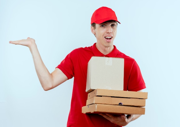 Молодой курьер в красной форме и кепке, держащий коробку с пакетом и коробки для пиццы, представляя что-то рукой, улыбаясь, стоя над белой стеной