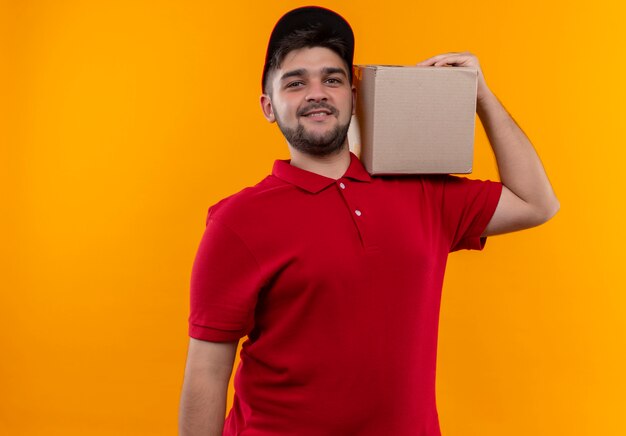 Young delivery man in red uniform and cap holding box package on shoulder looking confident