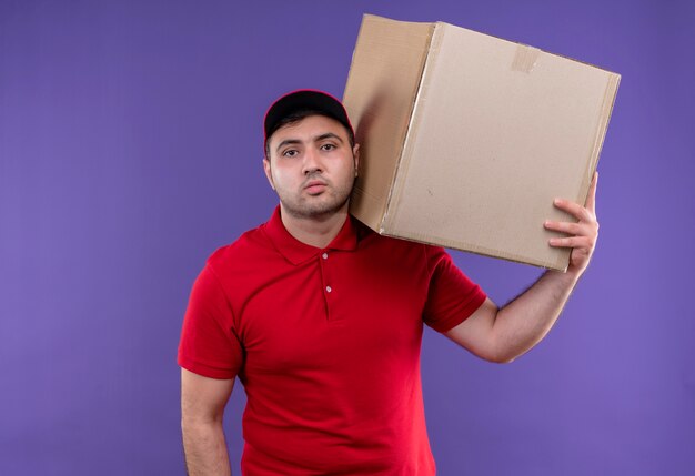 Young delivery man in red uniform and cap holding box package on shoulder with confident expression standing over purple wall