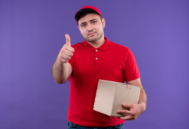 Young delivery man in red uniform and cap holding box package smiling confident showing thumbs up standing over purple wall