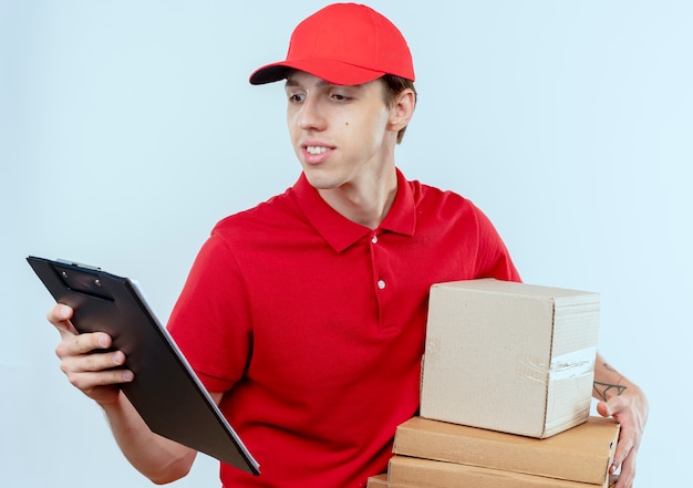 Young delivery man in red uniform and cap holding box packages and clipboard looking confident standing over white wall