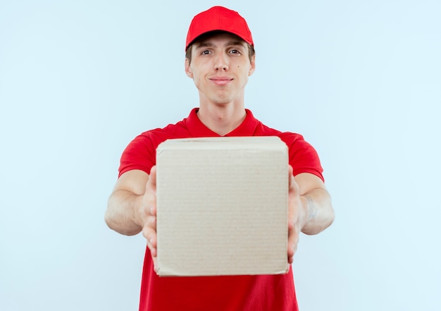 빨간 제복을 입은 젊은 배달 남자와 흰 벽 위에 서있는 자신감있는 표정으로 정면을 바라 보는 골판지 상자를 들고 모자