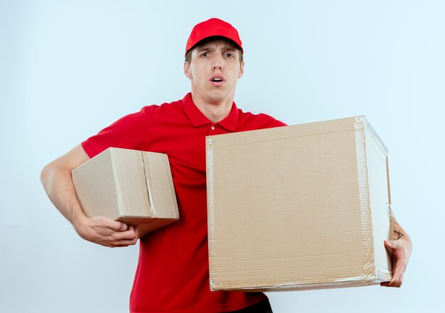 Young delivery man in red uniform and cap holding cardboard boxes looking to the front confused and surprised standing over white wall