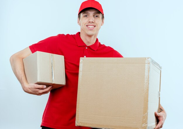 Young delivery man in red uniform and cap holding cardboard boxes looking to the front smiling confident standing over white wall