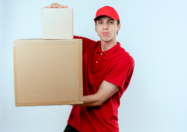 Young delivery man in red uniform and cap holding cardboard boxes looking to the front with serious expression standing over white wall