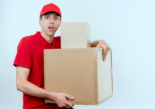 Young delivery man in red uniform and cap holding cardboard boxes looking to the front worried with fear expression standing over white wall