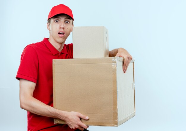 빨간 제복을 입은 젊은 배달 남자와 흰 벽 위에 서있는 두려움 식으로 걱정되는 전면을보고 골판지 상자를 들고 모자
