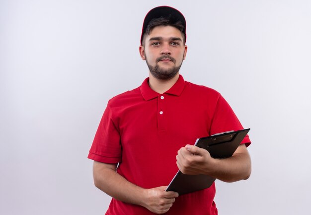 Young delivery man in red uniform and cap holding clipboard looking at camera with serious expression 