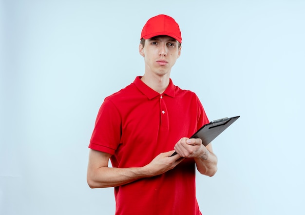 Giovane fattorino in uniforme rossa e cappuccio che tiene appunti guardando in avanti con la faccia seria in piedi sopra il muro bianco