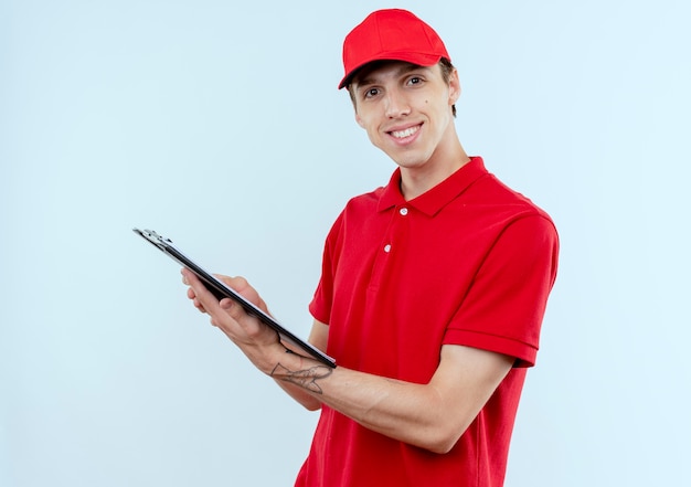 빨간 제복을 입은 젊은 배달 남자와 빈 페이지가 흰 벽 위에 자신감이 서있는 웃는 앞을 찾고 클립 보드를 들고 모자