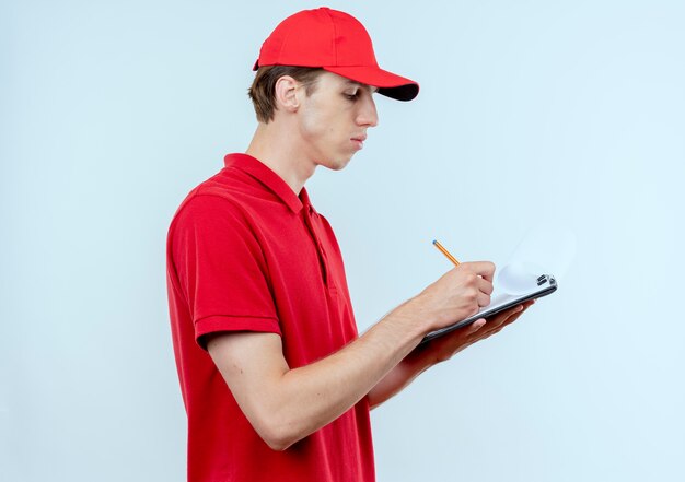 Young delivery man in red uniform and cap holding clipboard writing something with serious face standing over white wall 2