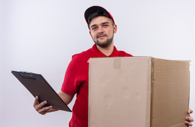 顔に笑顔で自信を持って見える大きな箱のパッケージとクリップボードを保持している赤い制服と帽子の若い配達人