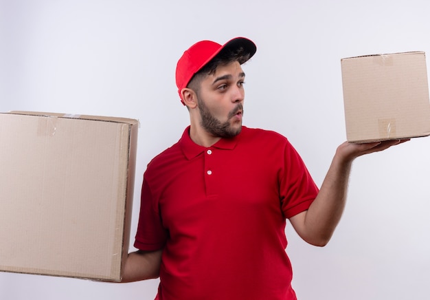 Молодой курьер в красной униформе и кепке держит большие картонные коробки, выглядит озадаченным, пытаясь сделать выбор