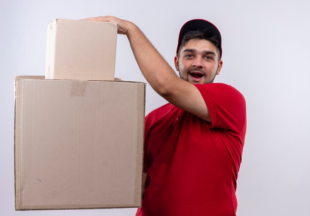 Young delivery man in red uniform and cap holding large cardboard boxes looking surprised and amazed 