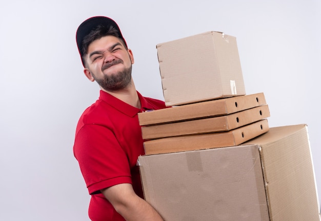 Young delivery man in red uniform and cap holding large cardboard boxes suffering from heavy weight 