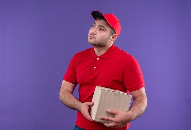 Молодой курьер в красной форме и кепке держит маленький пакет, глядя в сторону с уверенным выражением лица, стоя над фиолетовой стеной