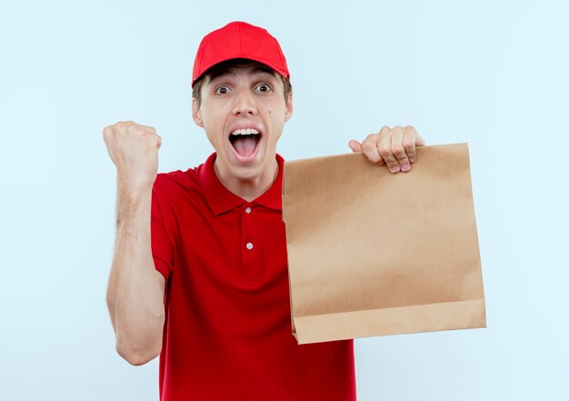 Молодой курьер в красной форме и кепке держит бумажный пакет, сжимая кулак, счастливый и взволнованный, стоя над белой стеной