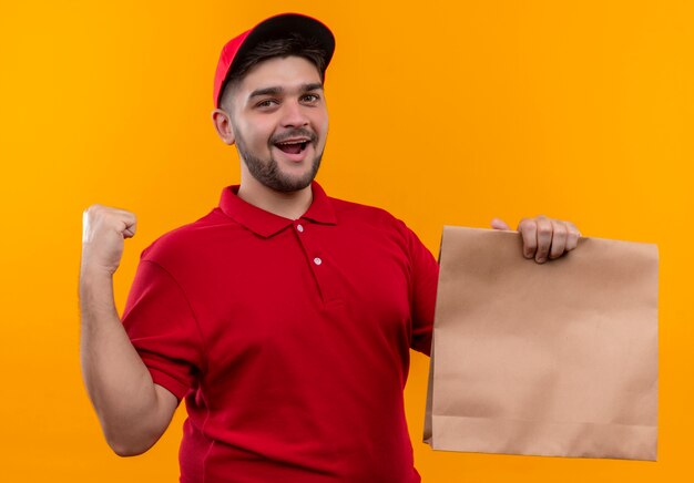 Young delivery man in red uniform and cap holding paper package clenching fist happy and exited smiling broadly