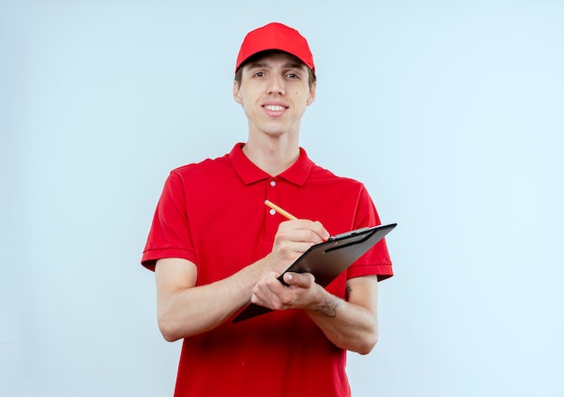 Молодой курьер в красной форме и кепке держит и карандаш смотрит вперед с уверенным выражением лица, стоя над белой стеной