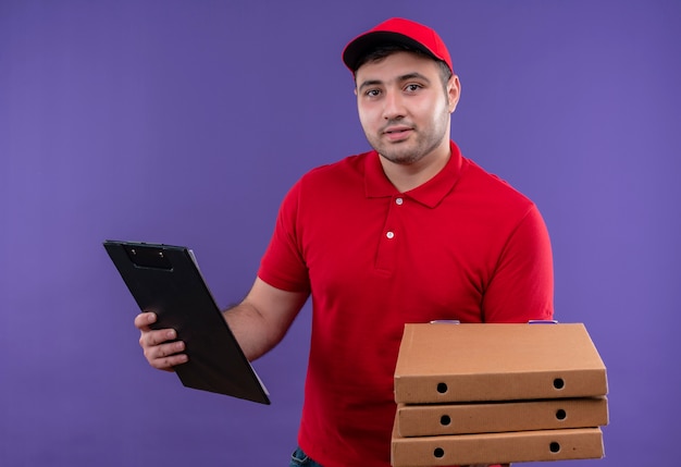 Young delivery man in red uniform and cap holding pizza boxes and clipboard smiling with happy face standing over purple wall