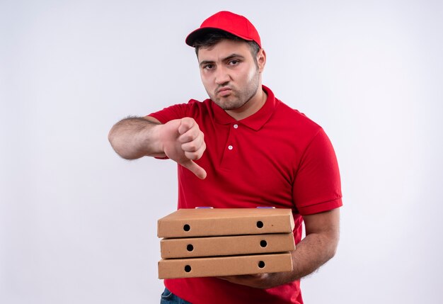 Молодой курьер в красной форме и кепке, держащий коробки с пиццей, недоволен, показывает палец вниз, стоя над белой стеной