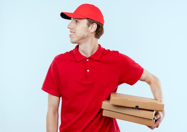 Молодой курьер в красной форме и кепке держит коробки для пиццы, глядя в сторону с уверенным выражением лица, стоя над белой стеной
