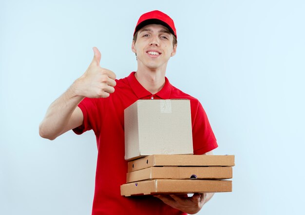 Молодой курьер в красной форме и кепке держит коробки для пиццы, глядя вперед, улыбаясь, уверенно показывая большие пальцы руки вверх, стоя над белой стеной