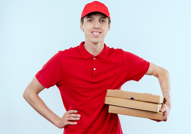 빨간 제복을 입은 젊은 배달 남자와 흰 벽 위에 서있는 자신감있는 표정으로 정면을 바라 보는 피자 상자를 들고 모자
