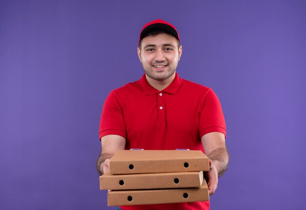 빨간 제복을 입은 젊은 배달 남자와 보라색 벽 위에 유쾌하게 서있는 피자 상자를 들고 모자