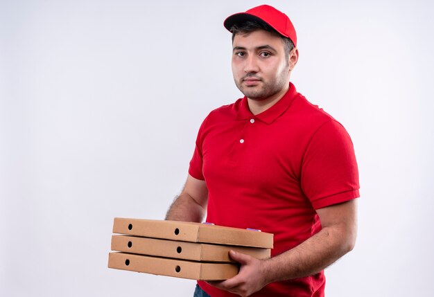 Молодой курьер в красной форме и кепке держит коробки для пиццы, улыбаясь с уверенным выражением лица, стоя над белой стеной