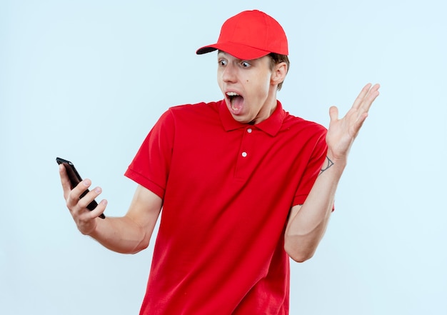 Giovane fattorino in uniforme rossa e cappuccio che tiene smartphone guardando sorpreso e confuso con il braccio alzato in piedi sopra il muro bianco