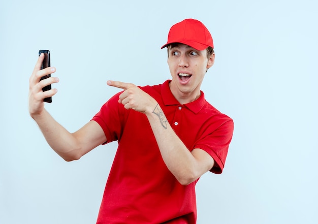 Молодой курьер в красной форме и кепке держит смартфон, принимая селфи, улыбаясь, указывая пальцем на камеру своего смартфона, стоящую над белой стеной