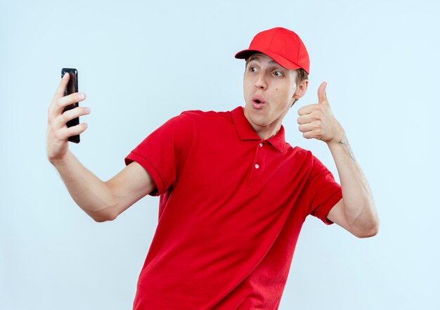 Young delivery man in red uniform and cap holding smartphone taking selfie smiling showing thumbs up standing over white wall