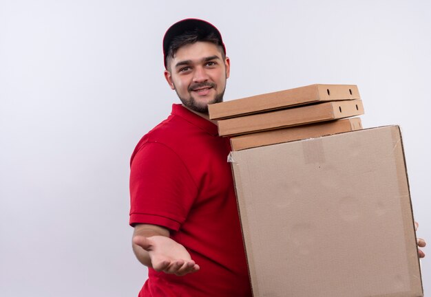 Young delivery man in red uniform and cap holding stack of boxes offering hand greeting smiling friendly 