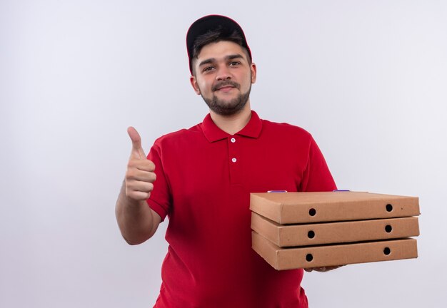 Young delivery man in red uniform and cap holding stack of pizza boxes looking confident smiling showing thumbs up 