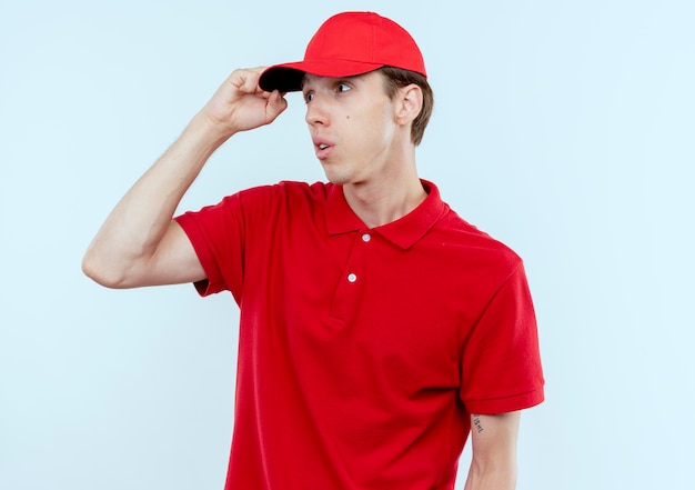 빨간 제복을 입은 젊은 배달 남자와 모자가 옆으로 손으로 혼란스러워 보임 오 흰 벽 위에 서있는 그의 머리