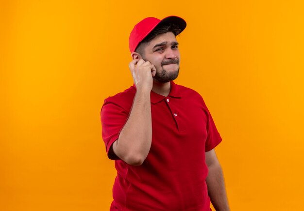 Young delivery man in red uniform and cap looking at camera with skeptic expression on face touching ear