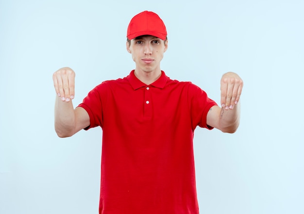 Giovane fattorino in uniforme rossa e berretto guardando fiducioso gesticolando con le mani, concetto di linguaggio del corpo in piedi sopra il muro bianco