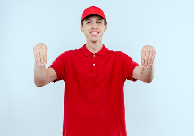 Young delivery man in red uniform and cap looking confident smiling gesturing with hands, body language concept standing over white wall