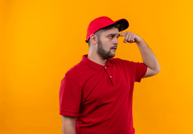 Young delivery man in red uniform and cap looking confused pointing his temple