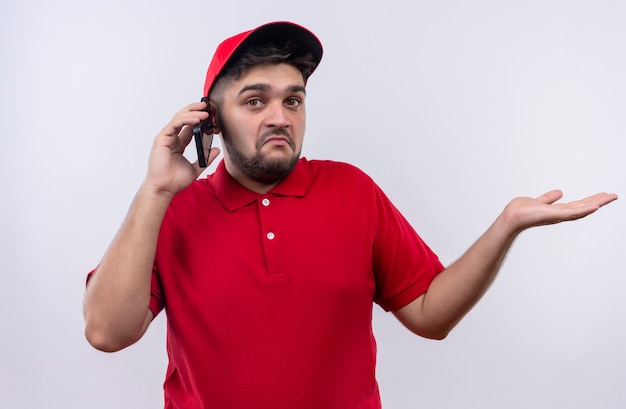 Молодой курьер в красной форме и кепке выглядит смущенным во время разговора по мобильному телефону