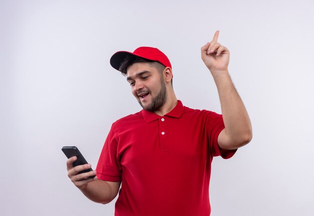 Молодой курьер в красной форме и кепке, глядя на экран своего смартфона, указывая пальцем вверх, имея отличную идею