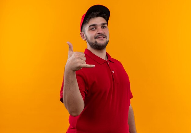 Young delivery man in red uniform and cap making call me gesture with smile on face