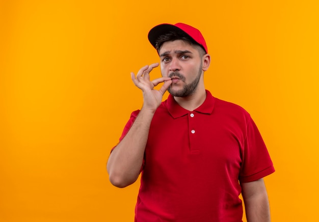 Young delivery man in red uniform and cap making silence gesture like closing mouth with a zipper