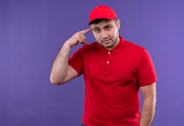 Young delivery man in red uniform and cap pointing with finger his temple looking confident standing over purple wall