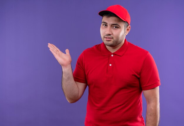 Young delivery man in red uniform and cap presenting copy space with arm of his hand standing over purple wall