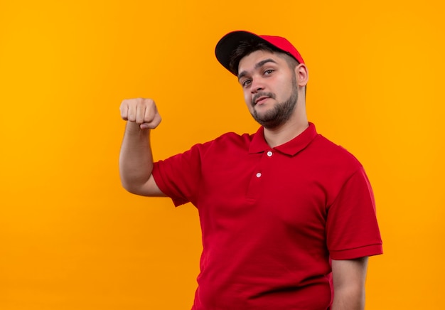 Giovane fattorino in uniforme rossa e cappuccio che alza il pugno sorridente amichevole