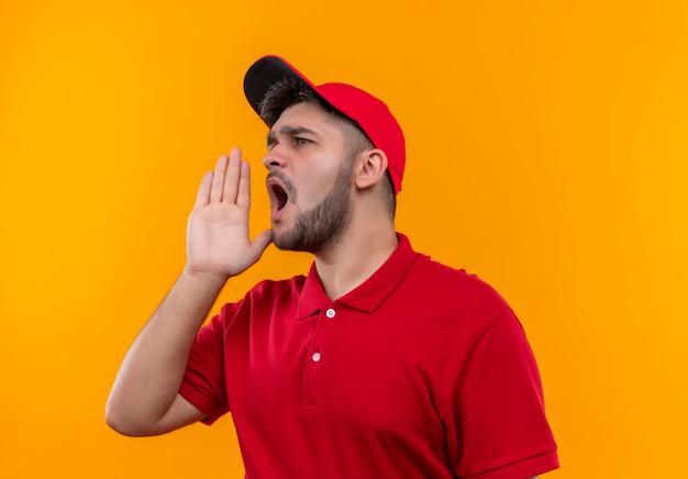 Young delivery man in red uniform and cap shouting or calling someone with hand near mouth