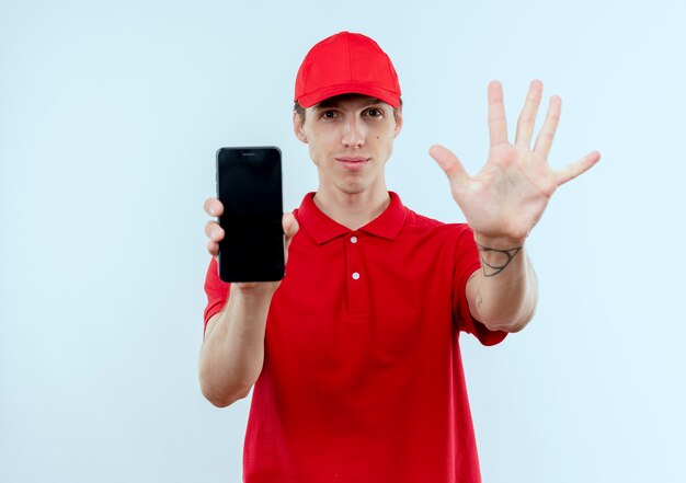 Young delivery man in red uniform and cap showing smartphone showing and pointing up with fingers number five smiling standing over white wall