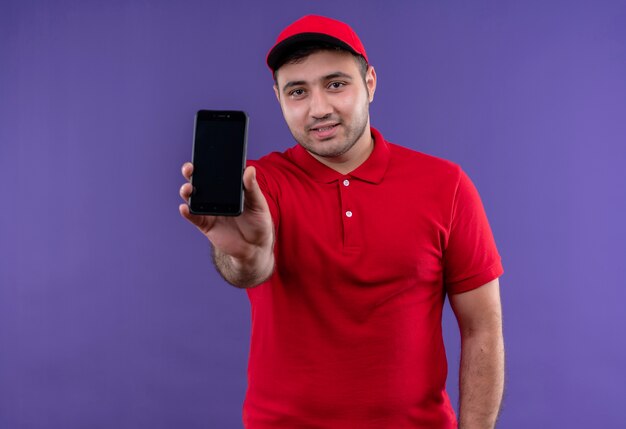 赤い制服とキャップの若い配達人は、紫色の壁の上に立って自信を持って笑顔のスマートフォンを示しています